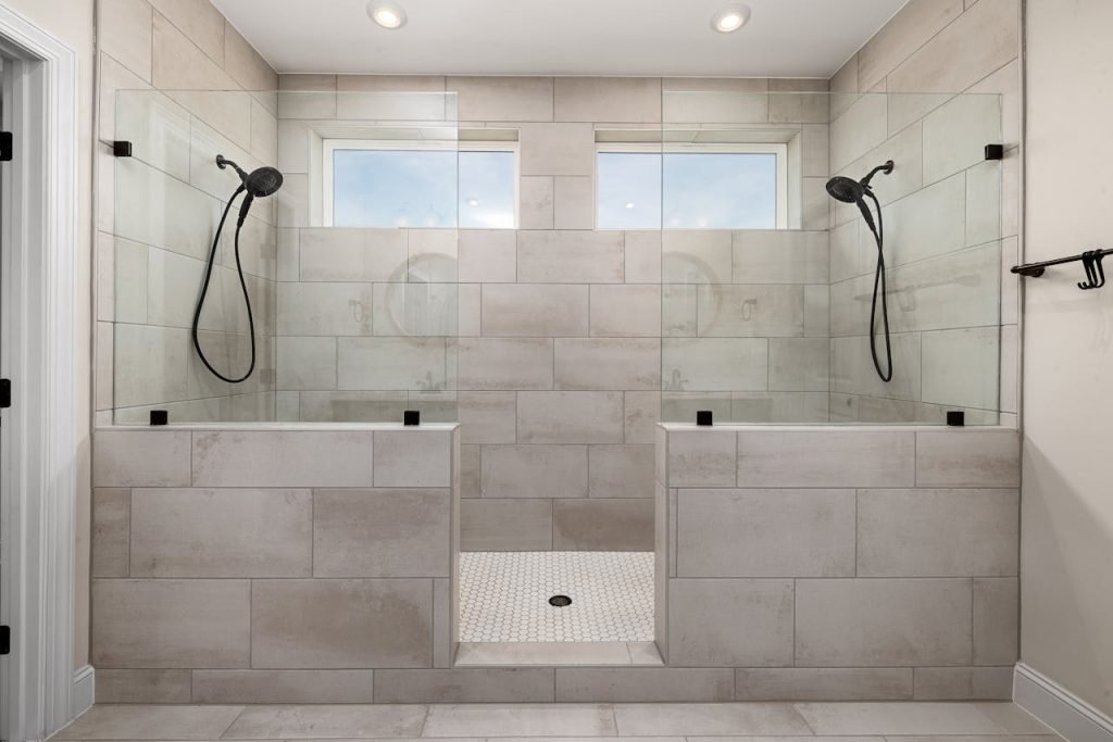 Modern dual walk-in shower with brushed nickel showerheads, glass partitions, and neutral tile walls under recessed ceiling lights.