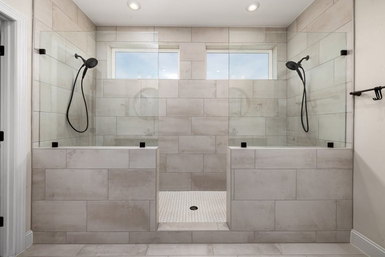 Modern dual walk-in shower with brushed nickel showerheads, glass partitions, and neutral tile walls under recessed ceiling lights.